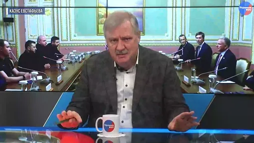Moderátor v televíznom štúdiu pred obrazovkou s prebiehajúcim rokovaním.