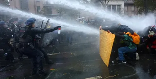 Policajný zásah vodným delom proti demonštrantom chrániacim sa drevenou doskou.
