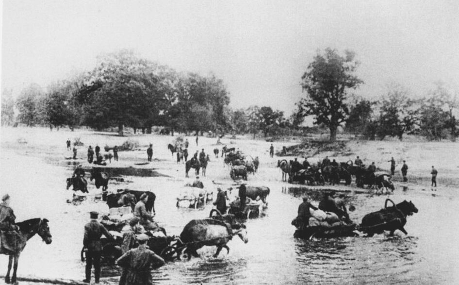 Historický konvoj s koňmi a vozmi brodiaci sa cez rieku.